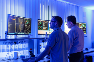 In einem in blaues Licht gehüllten Raum stehen zwei Männer vor drei Bildschirmen, auf denen Grafiken und technische Anzeigen zu erkennen sind. Die beiden Männer schauen konzentriert auf die Bildschirme, vor ihnen befinden sich Tastaturen und Computermäuse