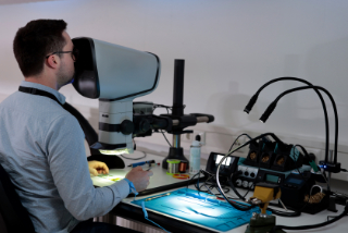 Von diagonal hinten ist ein Mann zu erkennen, der mit einem Mikroskop Computerchips untersucht. Rechts von dem Mikroskop befinden sich weitere technische Gerätschaften.
